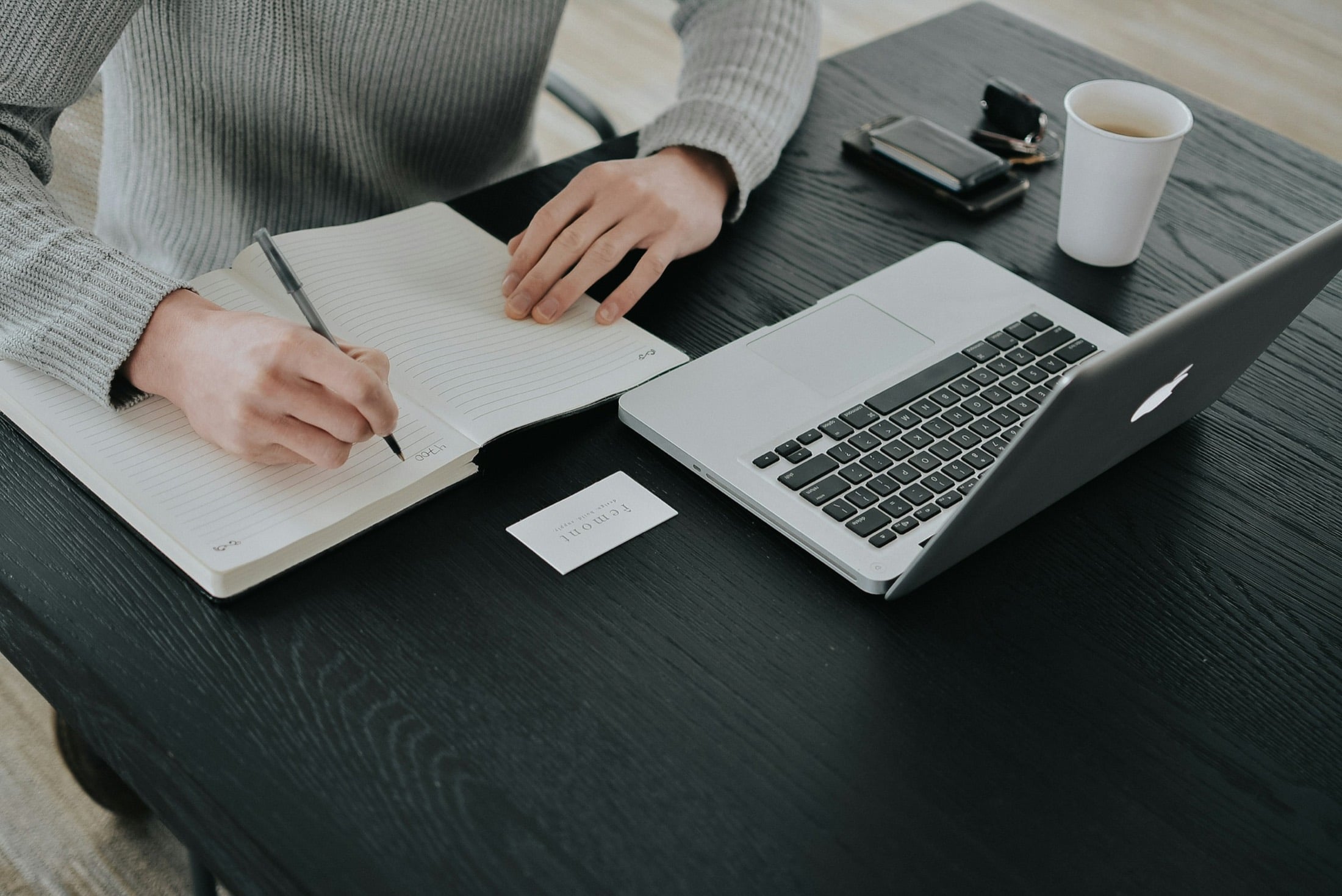 Hand writing notes in a notebook beside an open laptop