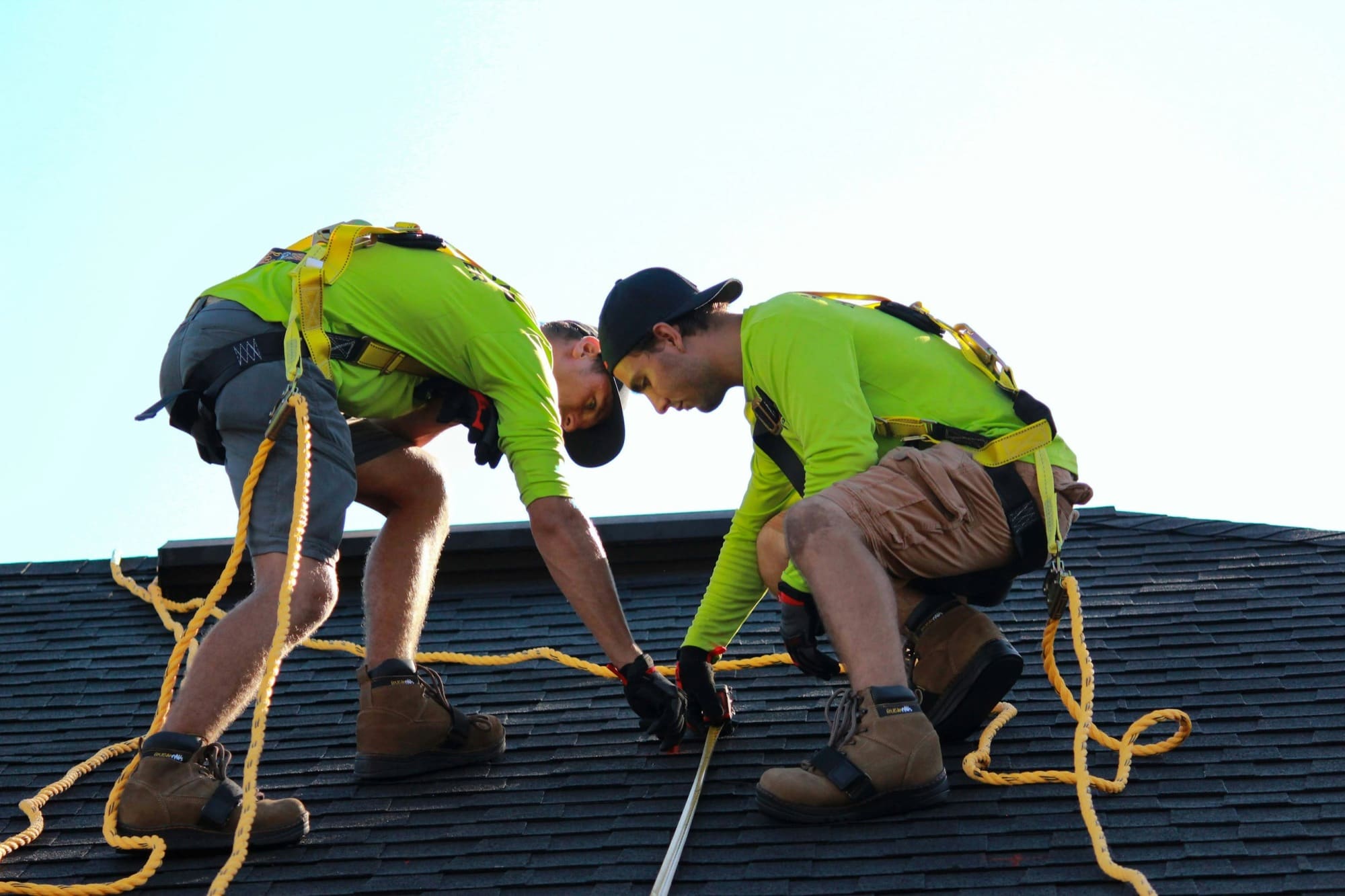 Two roofers in safety harnesses working closely together on a shingle roof